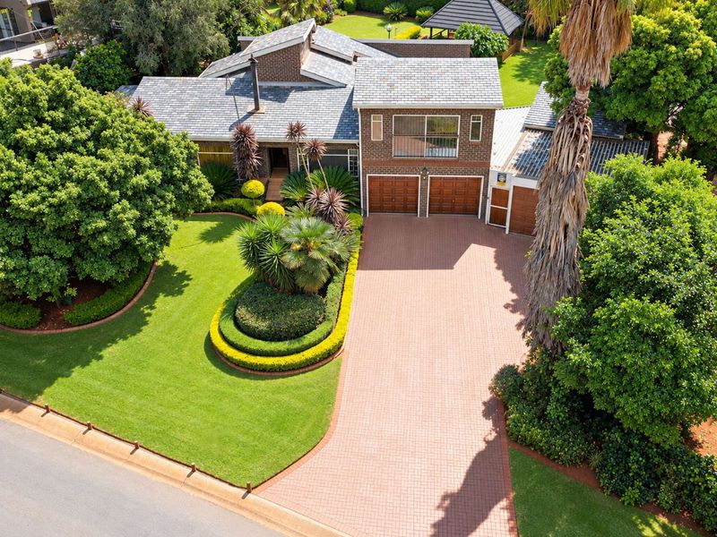 Aerial view of a well-maintained brick house featuring a double garage, paved driveway, and established landscaped gardens.
