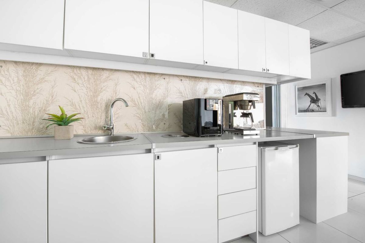 A compact office kitchenette featuring white cabinetry, a stainless steel sink, and a decorative glass splashback, providing essential utility for a professional workspace.