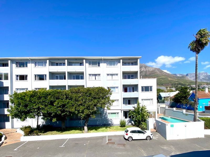 Exterior view of a multi-story apartment complex featuring balconies, a swimming pool, and an adjacent parking area with mountain views.