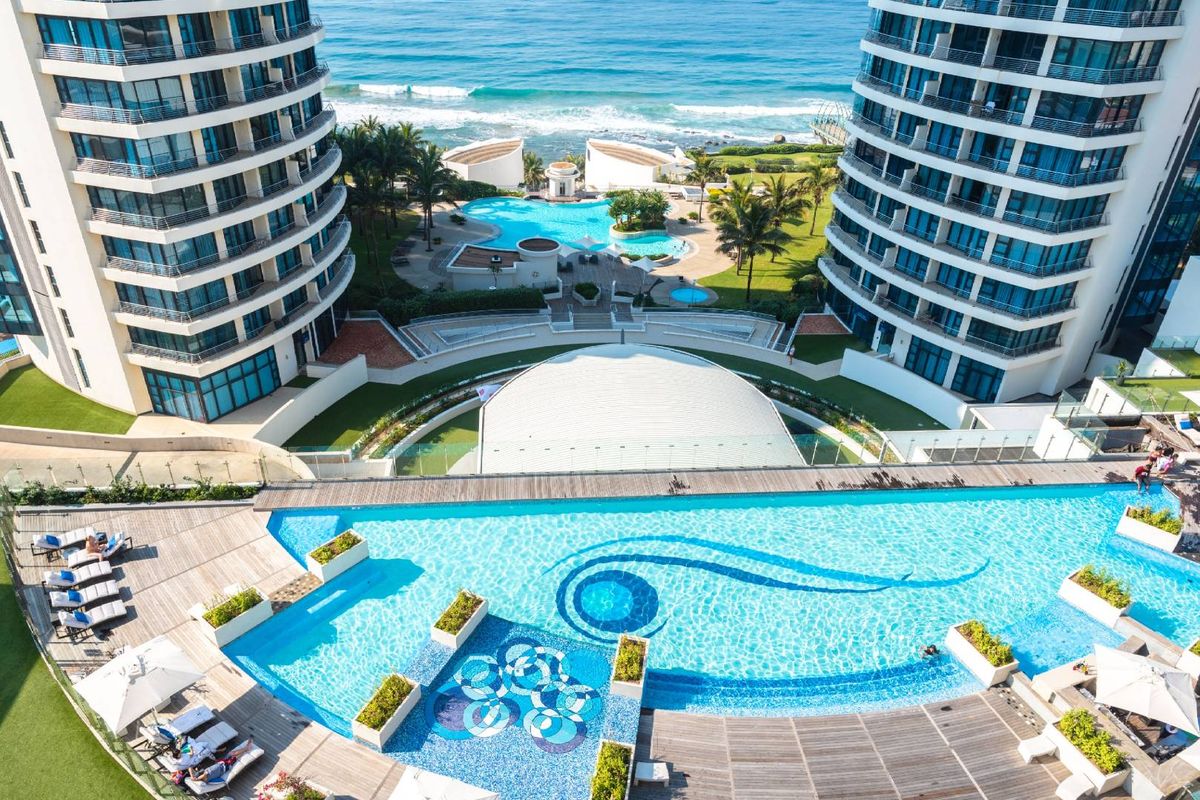 Aerial view of a luxury apartment complex featuring a large infinity swimming pool, landscaped grounds, and direct ocean views.