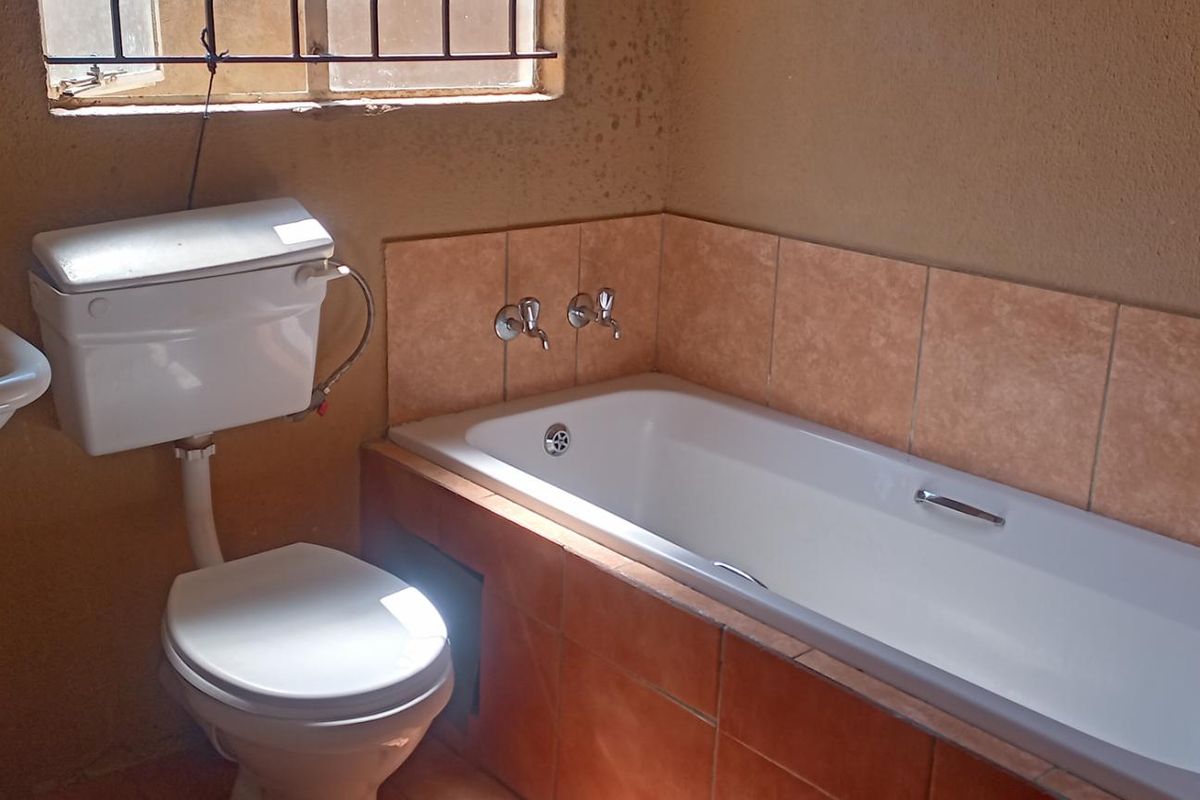 A functional bathroom featuring a standard bathtub, toilet, and wall-mounted basin with tiled splashbacks and security bars on the window.