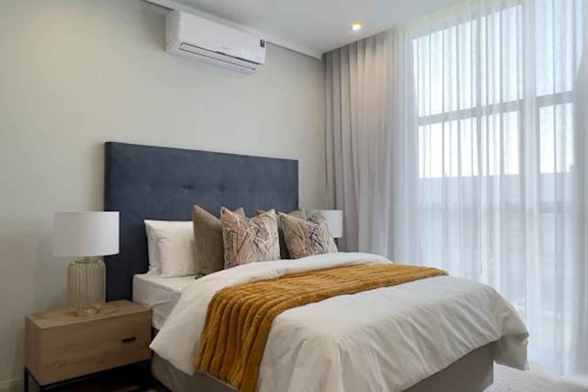 A modern, well-lit bedroom featuring neutral tones, split-system air conditioning, and floor-to-ceiling sheer curtains allowing for ample natural light.