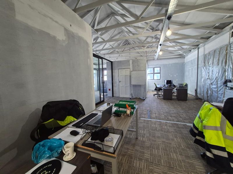 Open-plan office space featuring exposed roof trusses, carpeted flooring, and glass partitioning, currently undergoing renovation with unfinished wall surfaces.