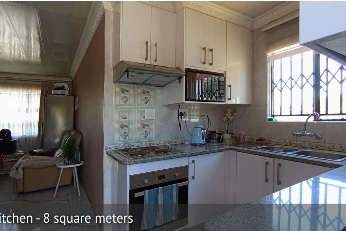 Compact kitchen featuring granite countertops, built-in oven, extractor fan, and white cabinetry, with an adjacent living area visible.