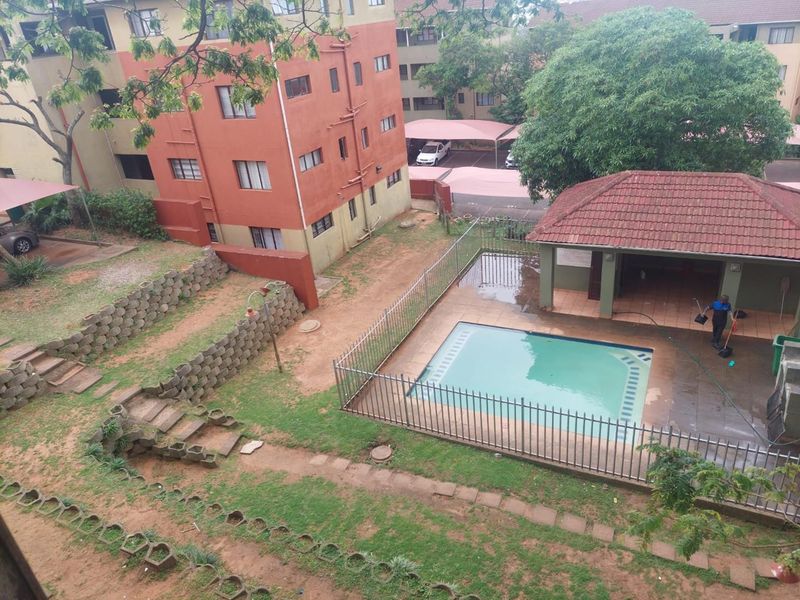 Aerial view of a communal apartment complex pool area featuring a fenced swimming pool, covered patio, and landscaped garden space.