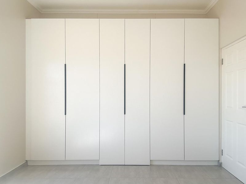 A clean, modern bedroom featuring floor-to-ceiling built-in cupboards with sleek black handles and neutral tiled flooring.