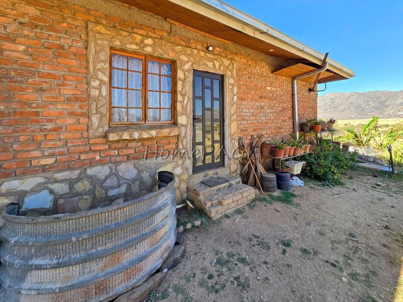 Rear exterior view of a rustic brick and stone cottage featuring timber-framed windows and a corrugated metal water tank in a rural setting.