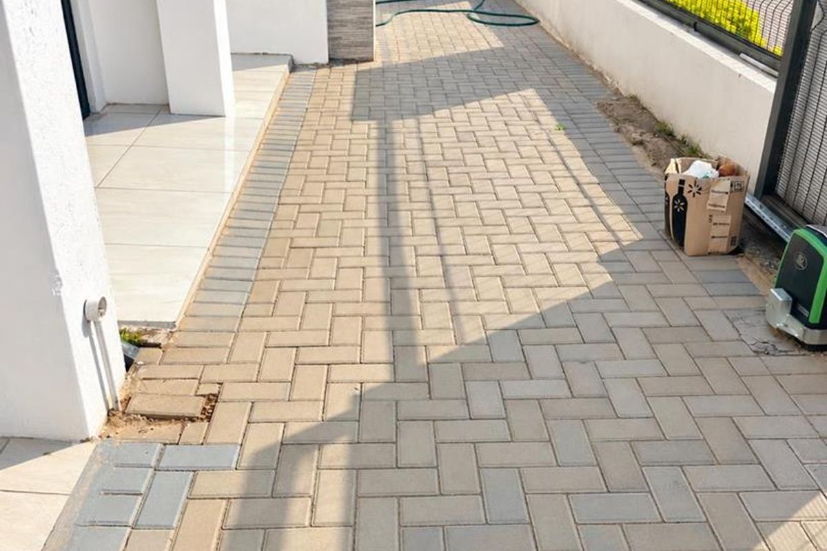 A paved side walkway or rear courtyard area featuring a boundary wall, metal gate, and adjacent tiled patio section.