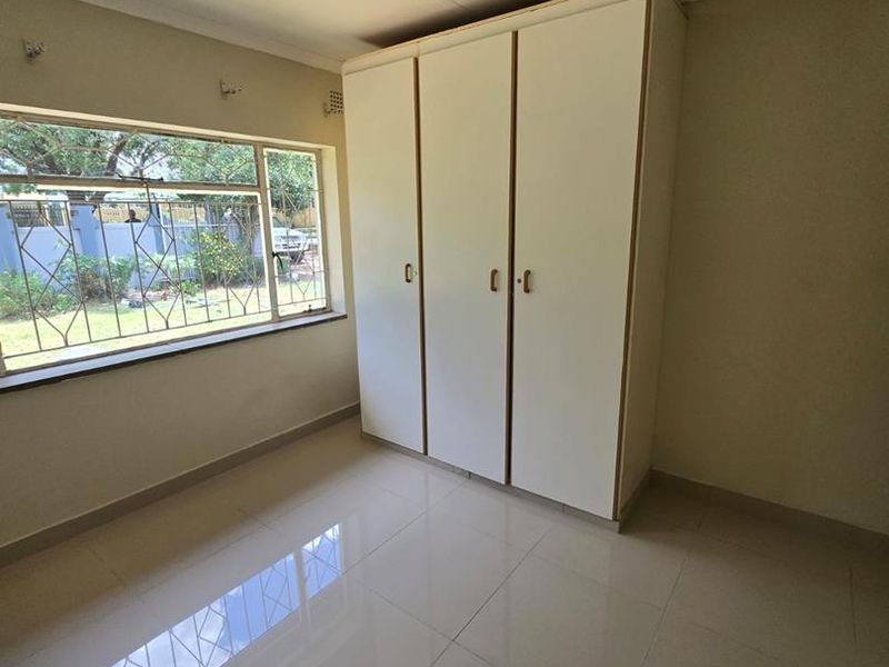 A bright, empty bedroom featuring tiled flooring, a built-in wardrobe, and large windows fitted with security bars.