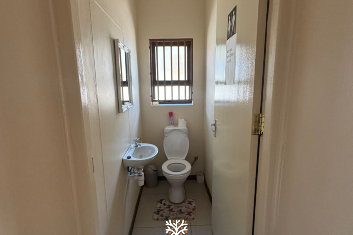 A compact office restroom featuring a wall-mounted basin, a standard toilet, and a small window with security bars.