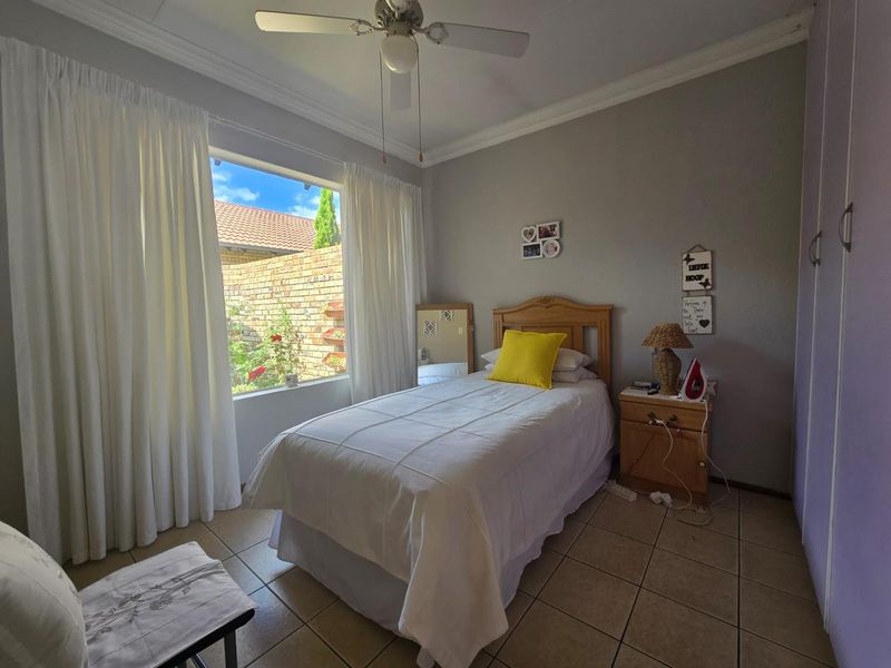 A well-lit bedroom featuring tiled flooring, built-in cupboards, and a ceiling fan, with a large window overlooking the rear garden area.