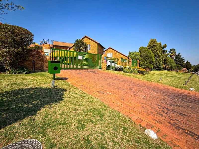 Front view of a townhouse property featuring a brick driveway, secure metal gate, and well-maintained lawn with established garden landscaping.