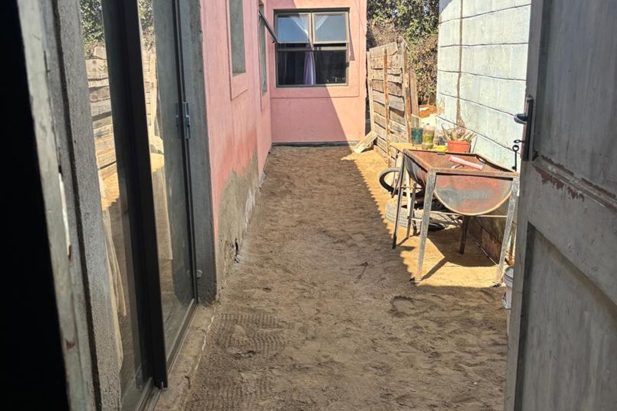 A narrow rear exterior passage featuring a sliding glass door, concrete block wall, and unpaved ground area requiring landscaping or paving.