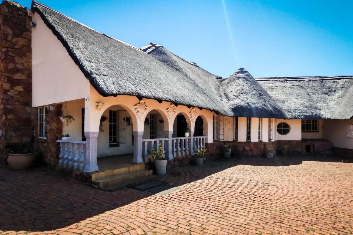 A charming farmhouse exterior featuring a traditional thatched roof, stone chimney, and a covered arched patio overlooking a brick-paved courtyard.