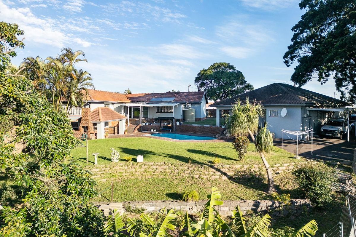Rear view of the property featuring a swimming pool, built-in braai area, landscaped garden, and paved driveway with ample space.