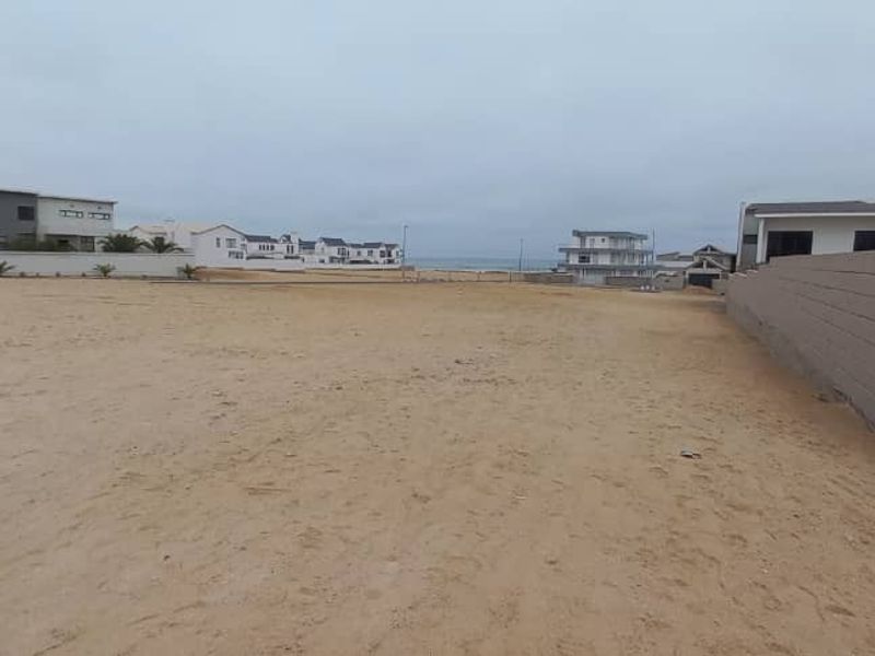 A vacant residential plot featuring sandy ground, a boundary wall, and a paved driveway, situated in a coastal neighborhood.