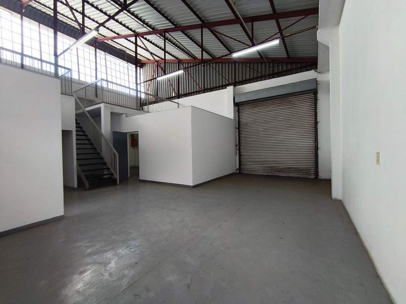 Spacious industrial warehouse unit featuring a high-clearance ceiling with exposed trusses, a mezzanine level, steel staircase, and a large roller shutter door.