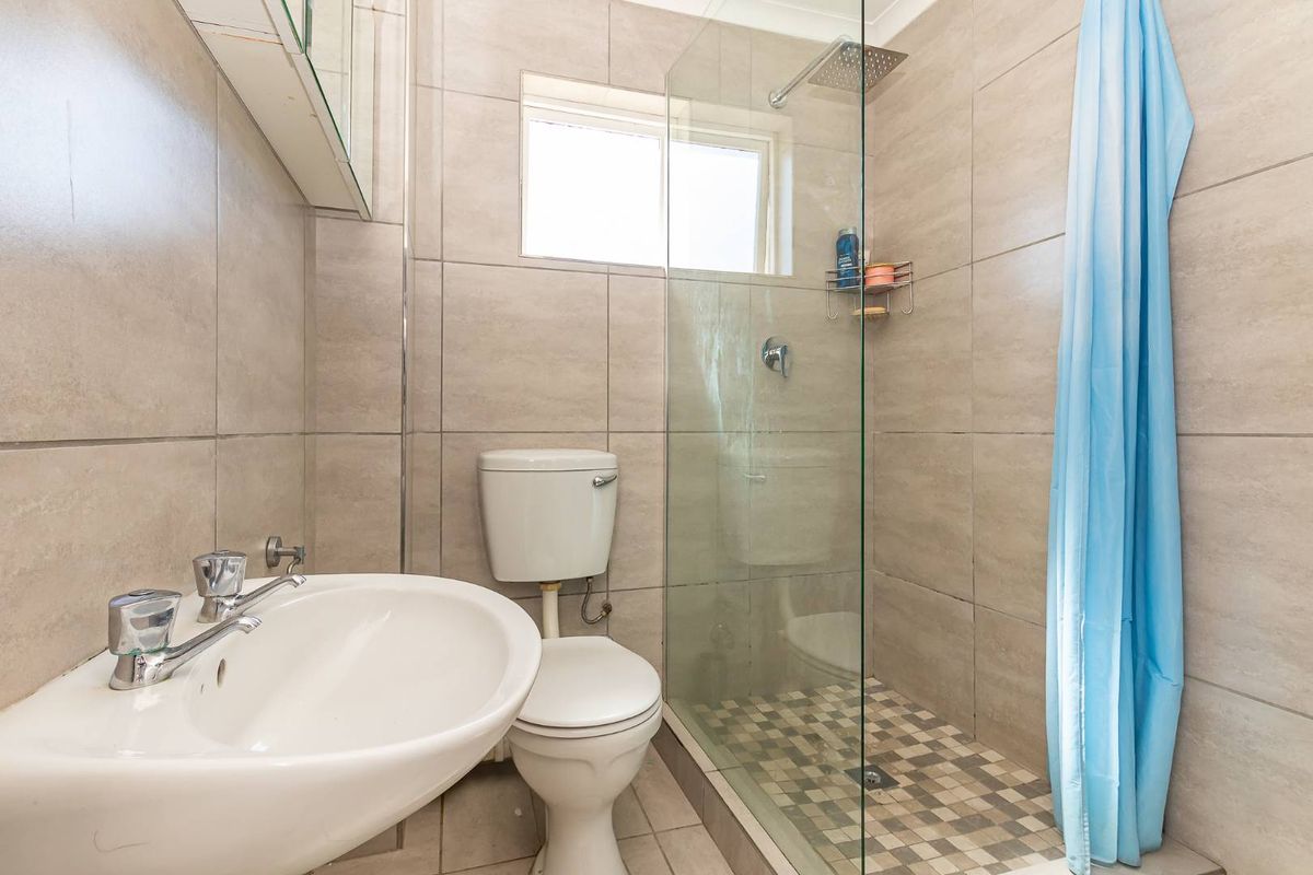 A compact bathroom featuring a glass-enclosed shower, pedestal basin, and toilet with neutral wall tiling and mosaic floor accents.
