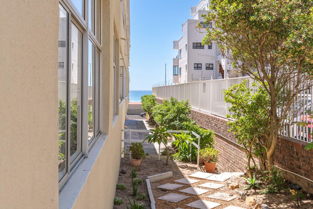 A side exterior view of the apartment building featuring a paved walkway, brick boundary wall, and security fencing with a glimpse of the ocean.