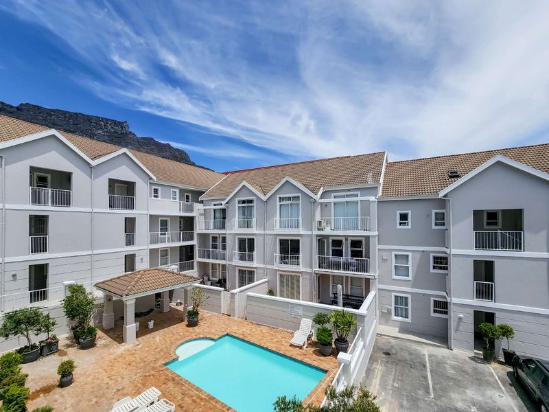 Aerial view of an apartment complex featuring a central swimming pool, paved courtyard, covered gazebo, and multiple residential balconies with mountain views.