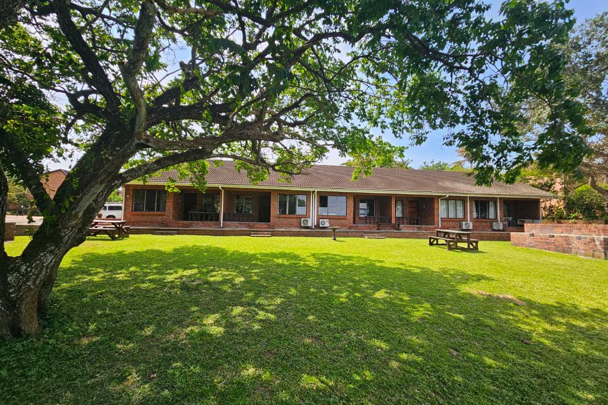 A spacious rear garden view of the lodge featuring a large lawn, mature trees, and a long brick building with multiple access points.