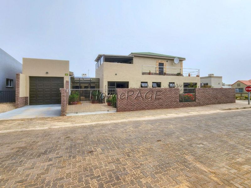 Front view of a two-story house featuring a brick boundary wall, metal gate, attached garage, and an upper-level balcony.