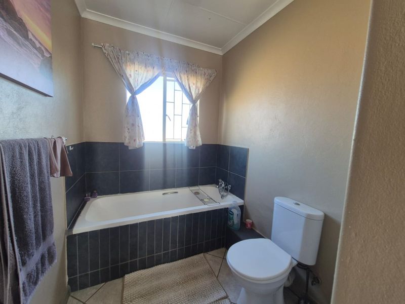 A functional bathroom featuring a built-in bathtub with dark tiled surround, a standard toilet, and a window with security bars.