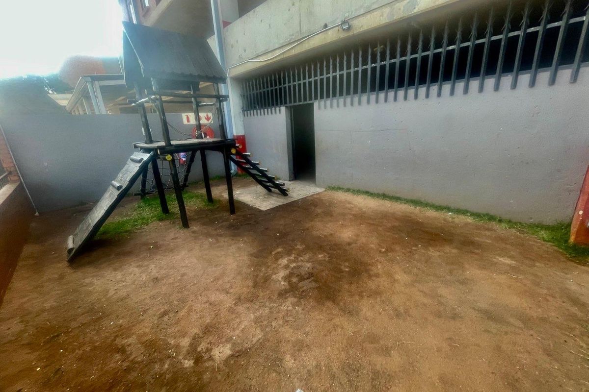 A communal outdoor area featuring a basic play structure, perimeter walling with security fencing, and an unlandscaped dirt ground surface.