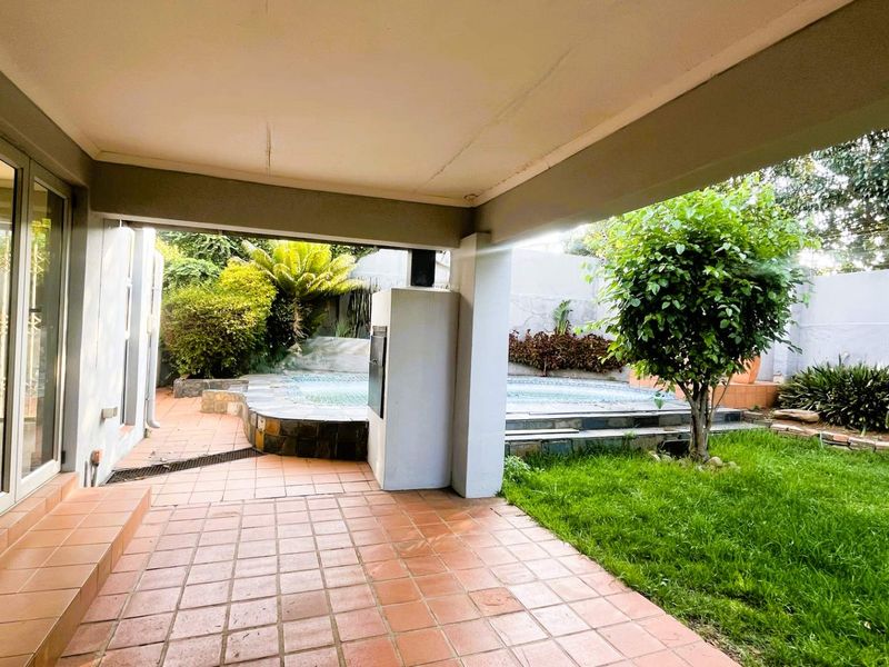 Covered patio area featuring tiled flooring and a built-in braai, overlooking a private swimming pool and landscaped garden space.