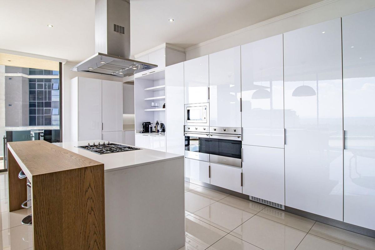 Modern open-plan kitchen featuring high-gloss white cabinetry, a central island with a gas hob, integrated stainless steel appliances, and sleek tiled flooring.