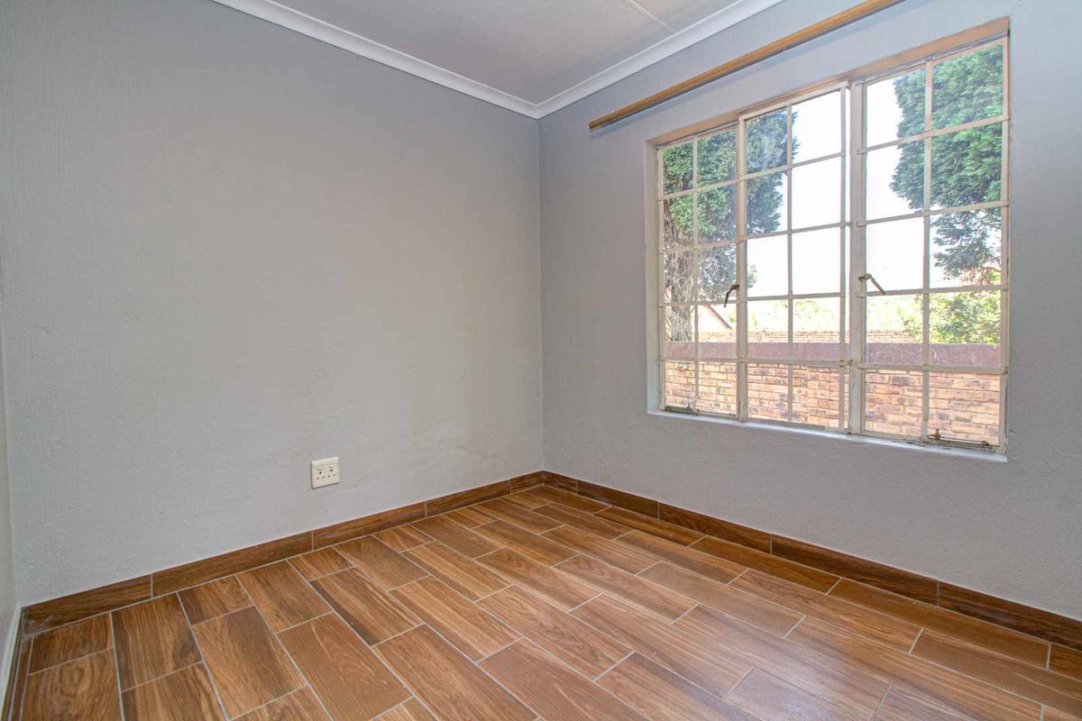 A bright, empty bedroom featuring wood-look tiled flooring, neutral grey walls, and a large window with metal burglar bars.