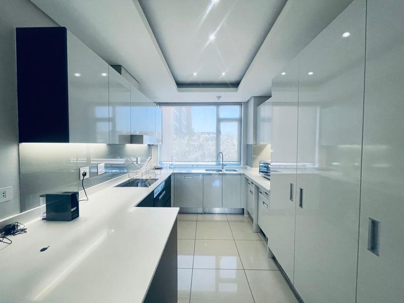Modern galley-style kitchen featuring high-gloss white cabinetry, quartz countertops, integrated appliances, and sleek tiled flooring with ample natural light.