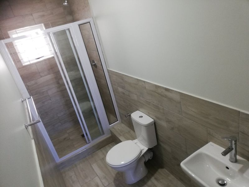 A modern, clean bathroom featuring a glass shower enclosure, white toilet, and wall-mounted washbasin with neutral wood-look wall and floor tiling.