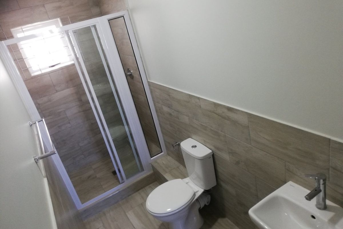 A modern, clean bathroom featuring a glass shower enclosure, white toilet, and wall-mounted washbasin with neutral wood-look wall and floor tiling.