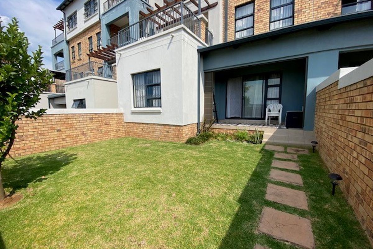 A private rear garden featuring a well-maintained lawn, paved pathway, and a covered patio area attached to the apartment building.