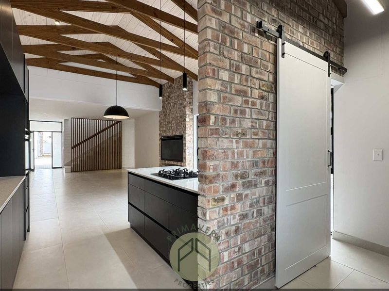 Modern open-plan living area featuring exposed timber beams, a striking brick feature wall, a sleek kitchen island with gas hob, and a sliding barn door.