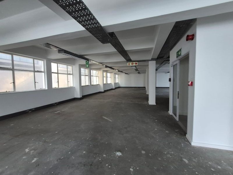 An expansive, open-plan office space featuring exposed ceiling cable trays, concrete flooring, and large windows providing ample natural light.