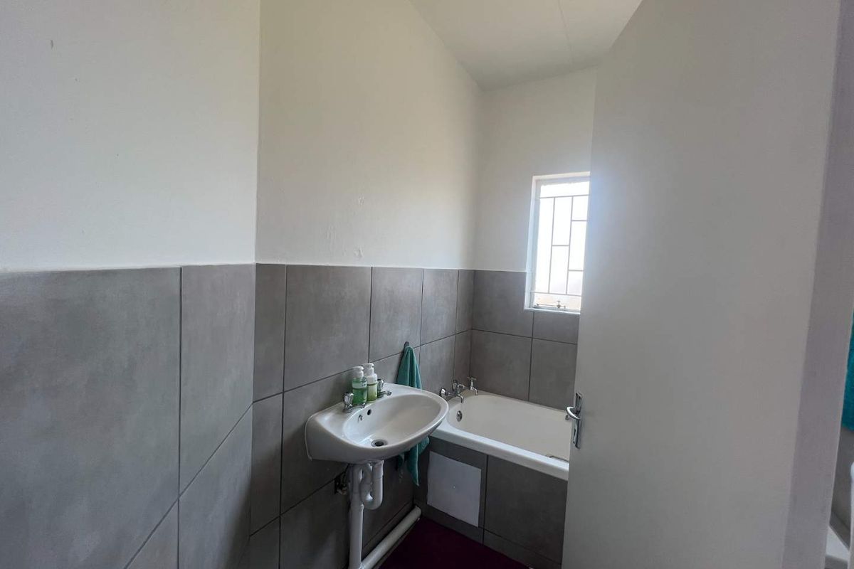 A functional bathroom featuring grey wall tiling, a built-in bathtub, and a wall-mounted basin with exposed plumbing.