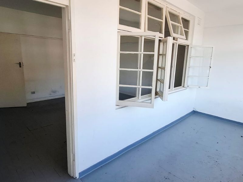 An empty, dated living area featuring painted concrete floors and multi-pane casement windows, requiring cosmetic updates to improve its overall condition.
