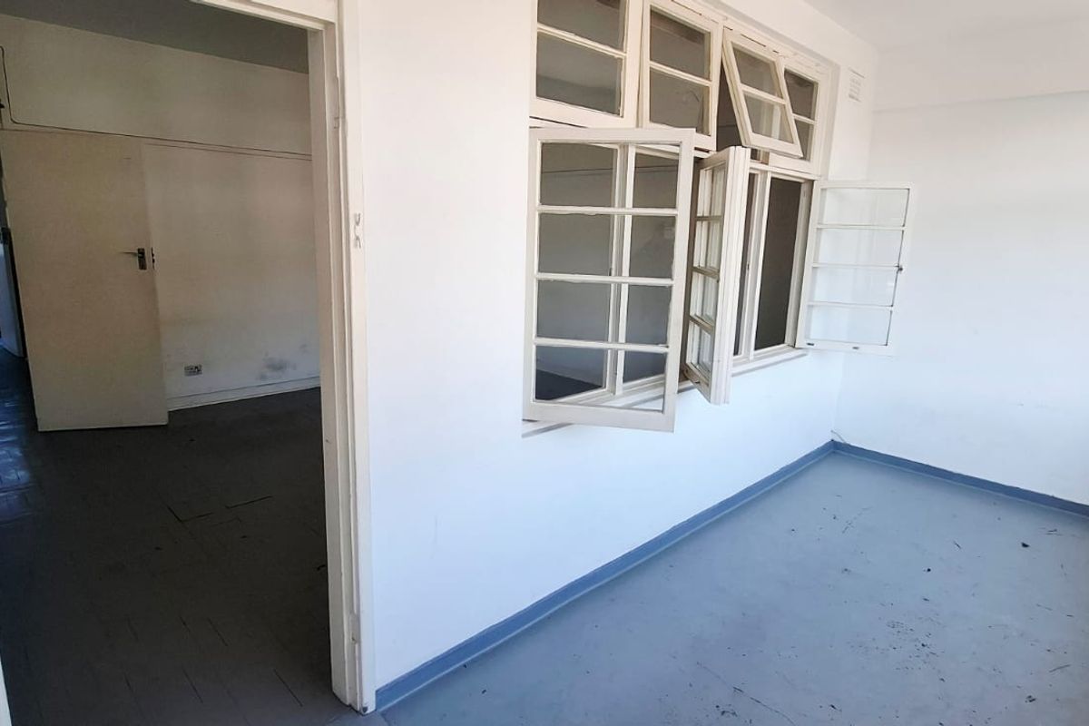 An empty, dated living area featuring painted concrete floors and multi-pane casement windows, requiring cosmetic updates to improve its overall condition.