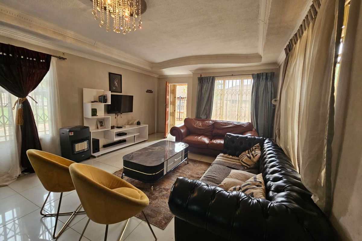 A spacious living room featuring tiled flooring, decorative ceiling molding, a crystal chandelier, and large windows with security bars.