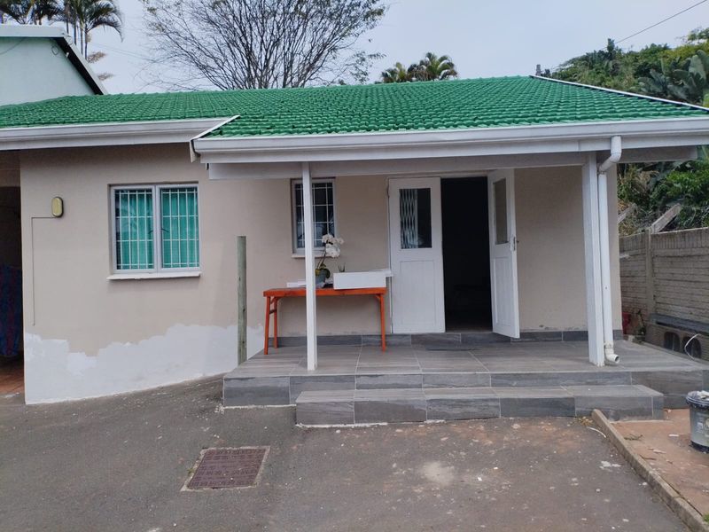 Front view of a cottage featuring a covered tiled patio, green metal roof, and security-fitted windows, with some exterior wall maintenance required.