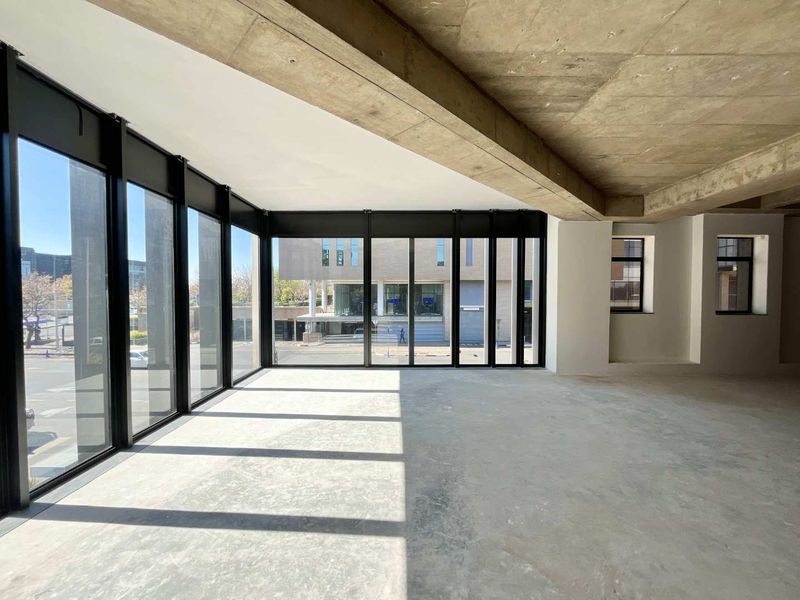 Spacious open-plan office area featuring exposed concrete ceilings, polished concrete floors, and expansive floor-to-ceiling windows providing ample natural light.