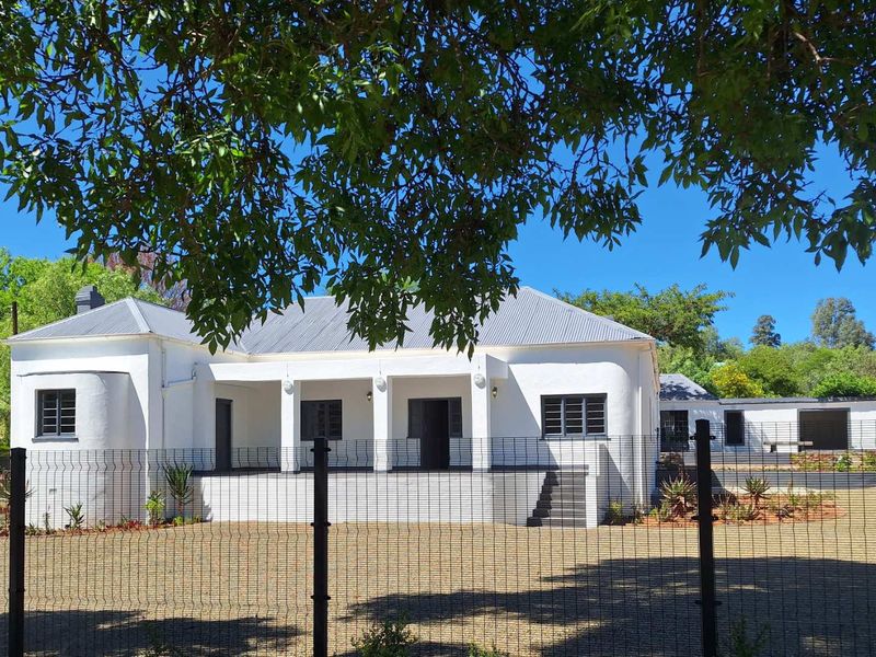 A well-maintained white guest house featuring a covered veranda, corrugated metal roof, and secure perimeter fencing set within a landscaped garden.