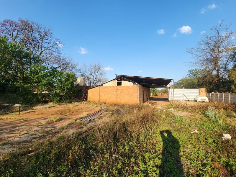 A view of a brick outbuilding with a metal roof, situated on a large, undeveloped plot with overgrown vegetation and perimeter fencing.
