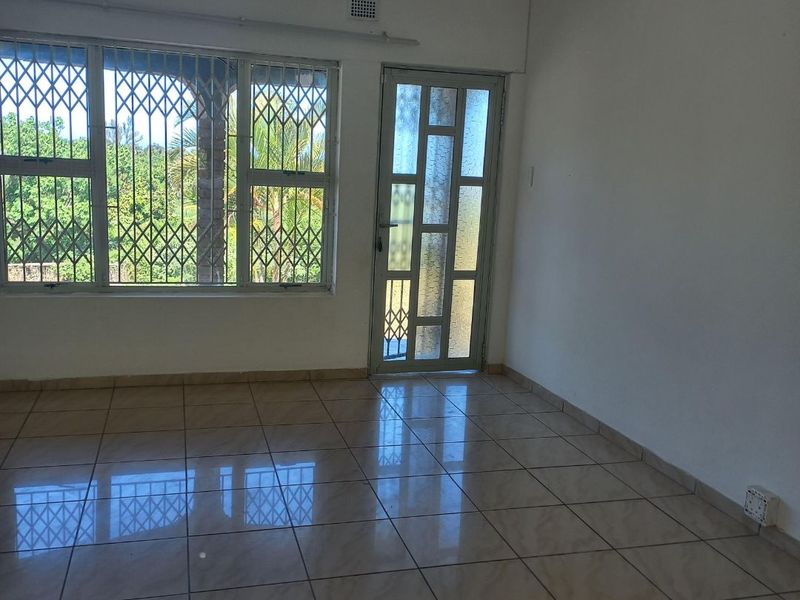 A bright, empty living area featuring neutral tiled flooring, large windows with security bars, and a glass-panelled door leading to the exterior.