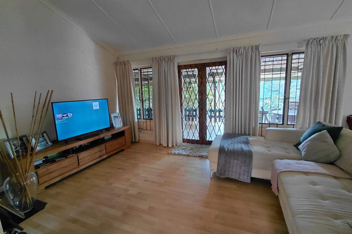 A comfortable living room featuring light wood-look flooring, large windows with curtains, and double doors providing access to the outdoor patio area.