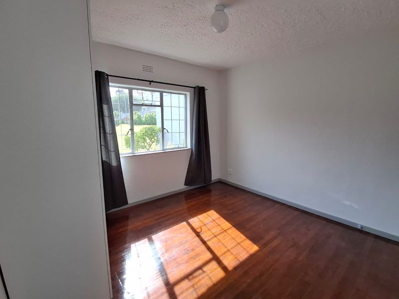 A bright, empty bedroom featuring polished parquet flooring, a built-in cupboard, and a large window allowing for ample natural light.