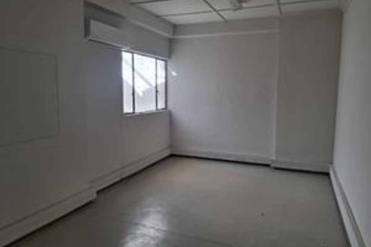 A vacant, neutral-toned office space featuring a suspended ceiling, wall-mounted air conditioning unit, and natural light from a multi-pane window.