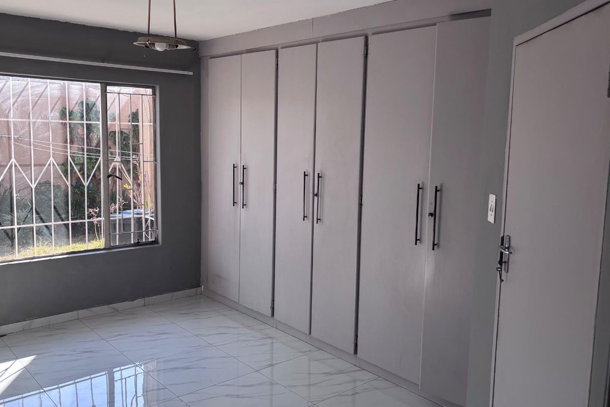 A bright, empty bedroom featuring modern tiled flooring, extensive built-in cupboards, and large windows fitted with security bars.